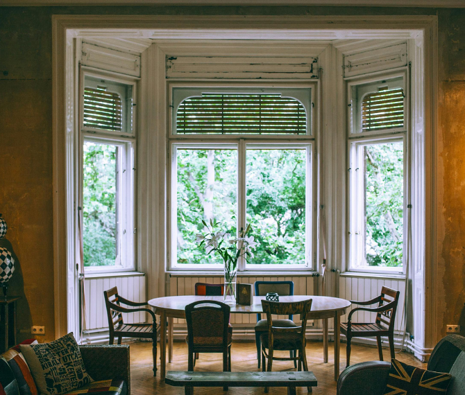 A large, open bay window with shutters and a table in the space with 6 chairs around it