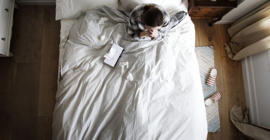 A woman sat in bed blowing her nose with a box of tissues and some used tissues next to her on the duvet.