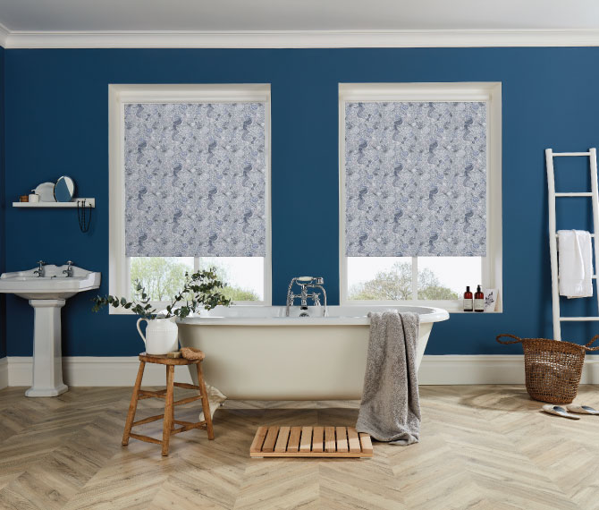 Roller Blinds A bathroom with a freestanding bath and dark blue walls. Centrally are two large windows with a blue patterned roller blind on each of them.